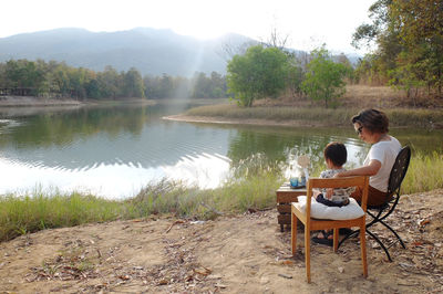 Woman and child sitting on chairs by lake against sky