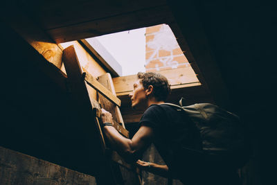 Side view of young man carrying backpack while climbing on ladder towards terrace