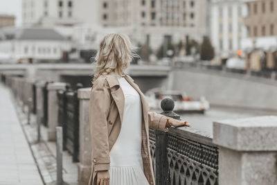 A middle-aged blonde woman with curly hair stands on the river embankment in the city.