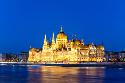 Illuminated hungarian parliament building by danube river against blue sky