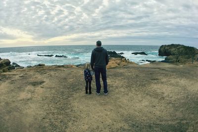 Full length rear view of father with daughter standing at sea shore against sky