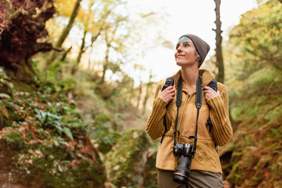 Low angle of traveling photographer standing in woods with photo camera and looking up while admiring nature during vacation