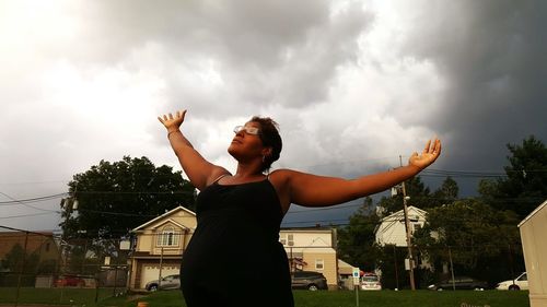 Woman with arms outstretched standing on field against cloudy sky