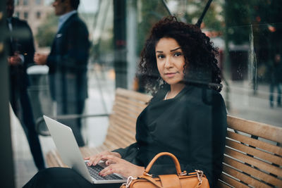 Portrait of mid adult businesswoman sitting with laptop at bus shelter seen from glass