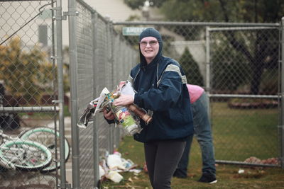 Portrait of happy woman cleaning garbage with friend by fence in yard