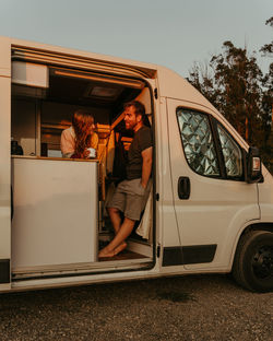 Cheerful couple of travelers standing in parked camper and looking at each other while enjoying sunset in summer during road trip in galicia