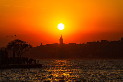 Silhouette buildings by sea against sky during sunset