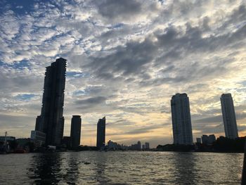 Modern buildings by river against sky during sunset