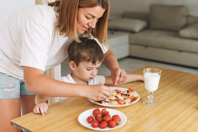 Cute toddler boy with mother having breakfast with puncakes and berries and glass of milk in kitchen 