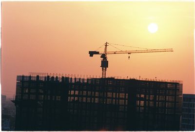 Silhouette crane by building against sky during sunset