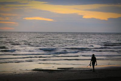 Silhouette of a fisherman walking on beach against sky during sunset