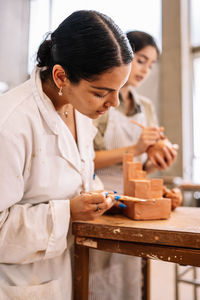 Side view merry hispanic girlfriends creating geometric shape from clay on table in spacious workshop