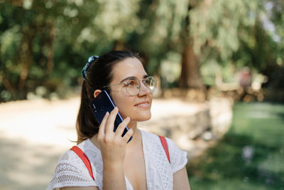 Medium shot of a young woman with a white dress and a red backpack talking on the phone in a park