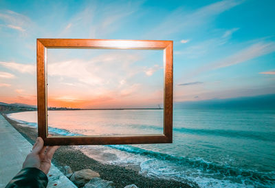 Cropped hand of person holding picture frame at beach during sunset