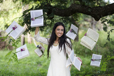 Digital composite image of smiling young woman holding book while standing in forest