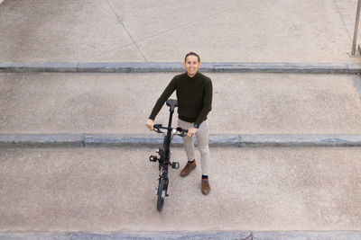 Top view of young man smiling to the camera going down the concrete steps with bicycle on his hands