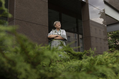 Real happy smiling senior woman in yard of modern building. green bushes around. copy space for text