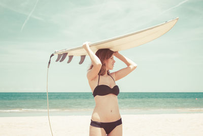 Young woman in bikini carrying surfboard on head while standing at beach against sky