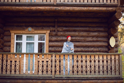 Woman in a red hat and scarf and a mug stands at a wooden house, in the woods in the autumn morning 