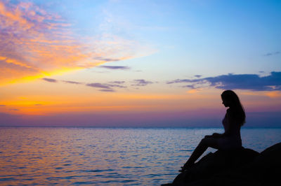 Woman sitting by sea against sky during sunset