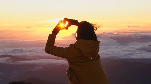 Rear view of woman making heart shape against sky during sunset