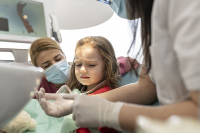 Dentist talking with cute little girl sitting by mother at medical clinic