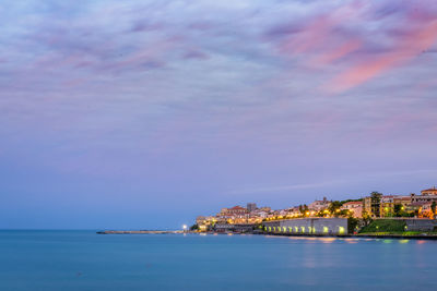 Buildings by sea against cloudy sky during sunrise