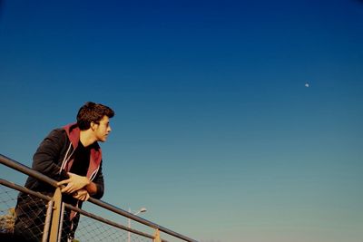 Low angle view of young man leaning on railing against clear sky