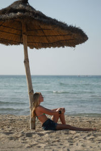 Side view of woman sitting at beach against sky