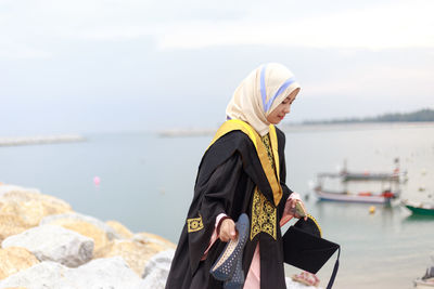 Young woman in graduation gown standing at beach against cloudy sky