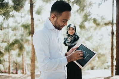 Side view of man writing on blackboard while woman standing in background