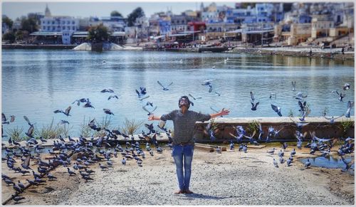 Full length of young man with arms outstretched standing amidst pigeons against lake in city