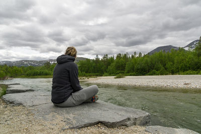 Woman sitting on rock by lake against sky