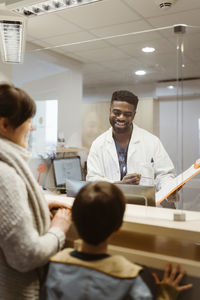 Smiling male receptionist talking to mother and son through transparent shield in clinic