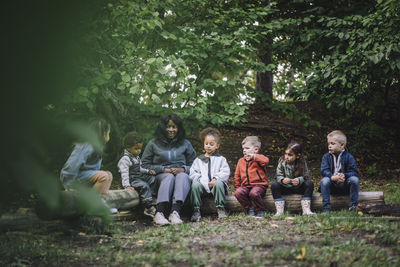 Female teacher sitting on log with preschool children in park