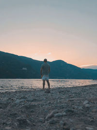 Rear view of man looking at mountain while standing by lake during sunset