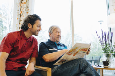 Smiling caregiver sitting by elderly man reading newspaper against window at retirement home