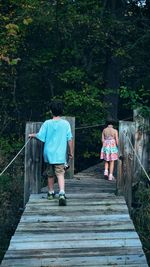 Rear view of girl and boy walking on wooden footbridge against trees in forest