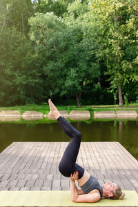 A slim woman on a wooden platform by a pond in a park in summer, does yoga