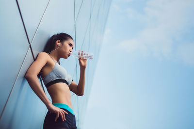 Low angle view of young woman drinking water from bottle while standing against wall