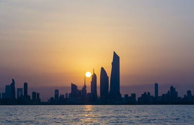 Sea by modern buildings against sky during sunset