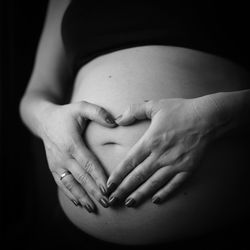 Midsection of pregnant woman making heart shape with hands on stomach against black background