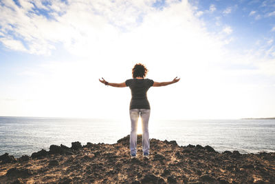 Rear view of woman standing with arms outstretched on rock at beach against sky