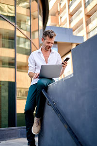 Low angle of mature businessman in smart casual clothes using laptop and smartphone while sitting on border during work on remote project on street