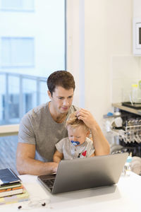 Baby boy sitting with father using laptop at table in house