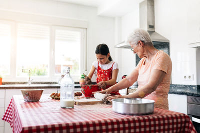 Cheerful grandma in eyewear against granddaughter with dough on hands during cooking process at table in house kitchen