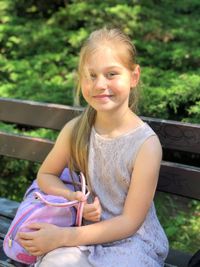 Portrait of smiling girl holding bag while sitting on bench at park