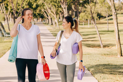 Two smiling young women walking in the morning park. carrying mats and other equipment for yoga.