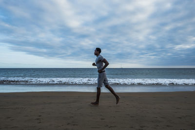 Full length of man running at beach against cloudy sky