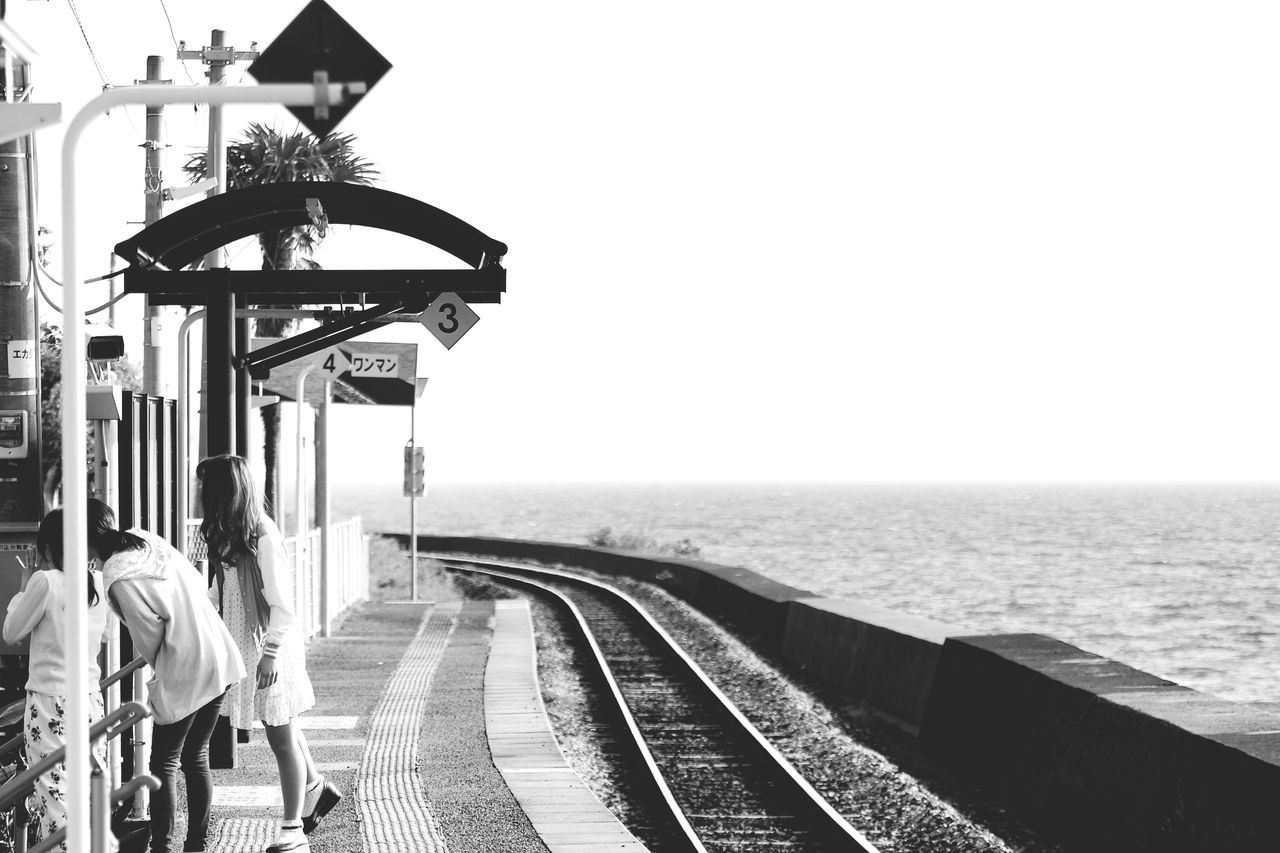 real people, transportation, clear sky, sea, railroad track, standing, travel, rear view, water, outdoors, lifestyles, waiting, men, public transportation, leisure activity, day, horizon over water, one person, full length, women, sky, nature, adult, people